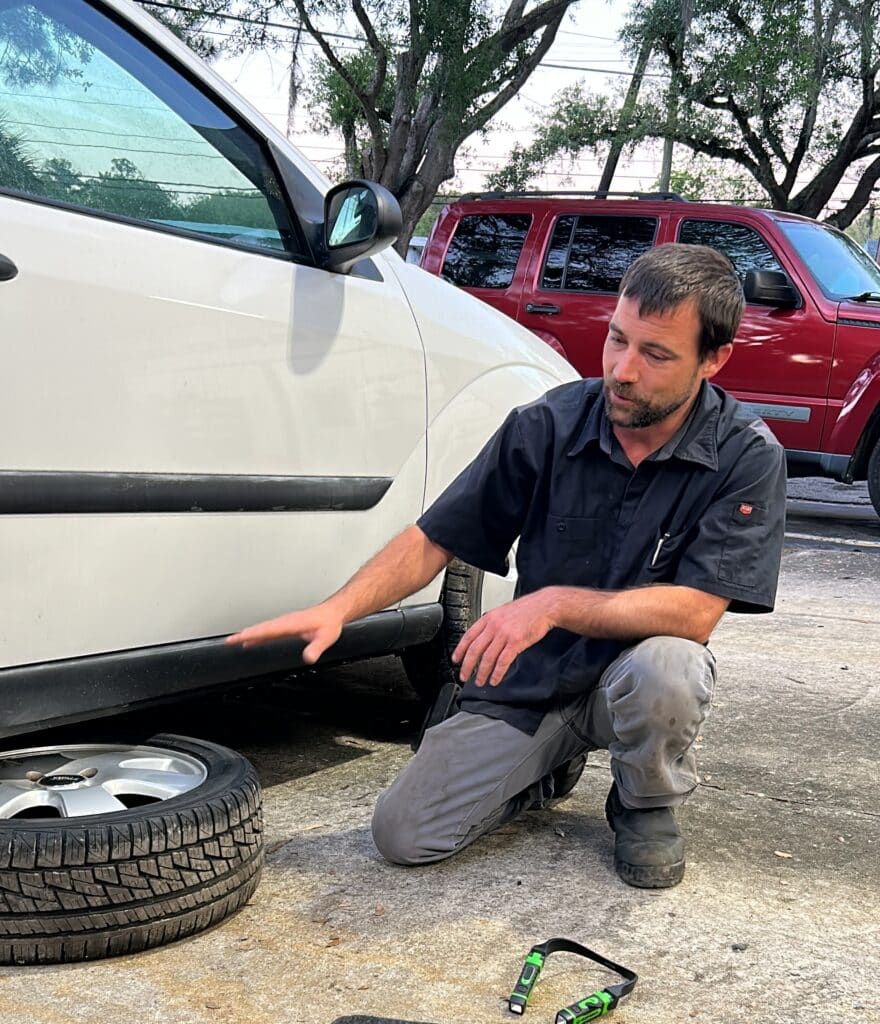 technician showing how to change a tire gestures to a flat tire placed underneath the car frame to prevent the car from hitting the ground in the event it slips off the jack. 