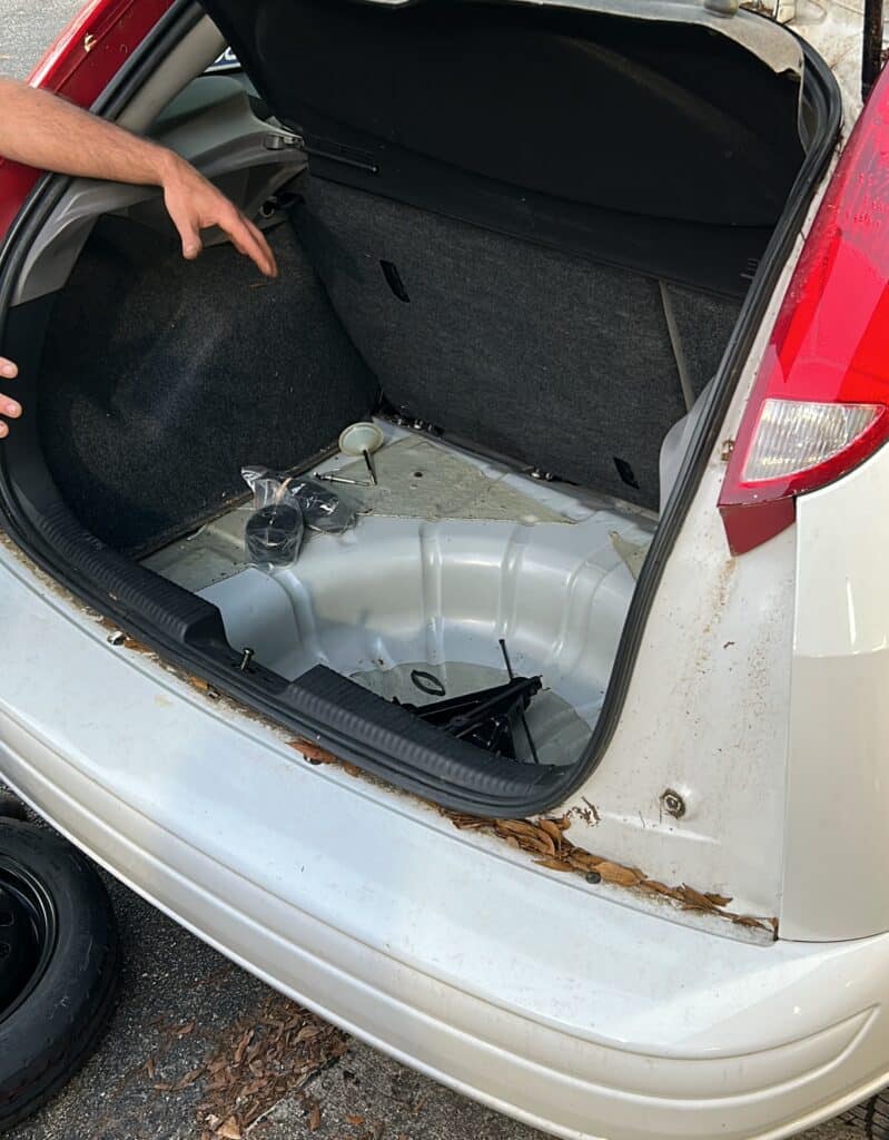 mechanic's hand gesturing to the location of a spare tire in the trunk of a small white car