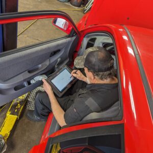 An auto technician sitting the the front seat of a red car completing a diagnostic test on the vehicle